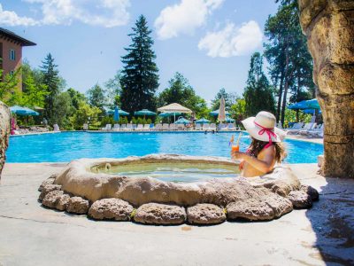 Жена по бански в джакузи пиеща коктейл и гледаща към минерален басейн в слънчев ден